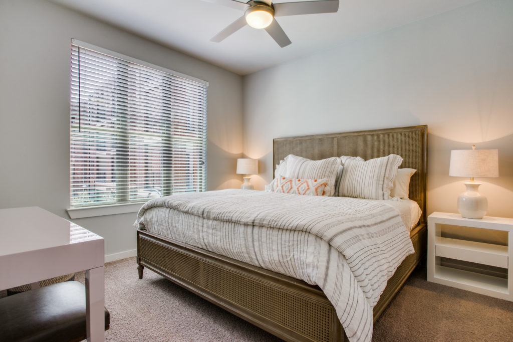 Carpeted apartment bedroom with queen bed, ceiling fan, and window with wood blinds