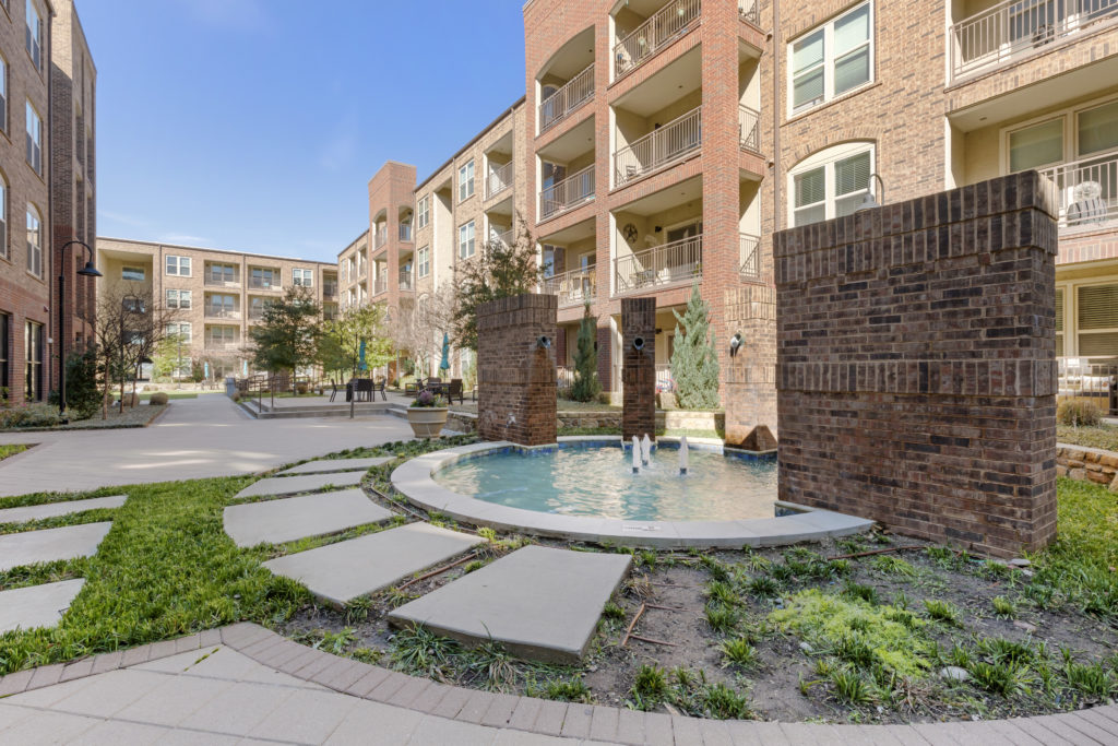 Outdoor fountain water feature with 3 water spouts, walkable paving and lush landscaping with apartment balconies in the background.