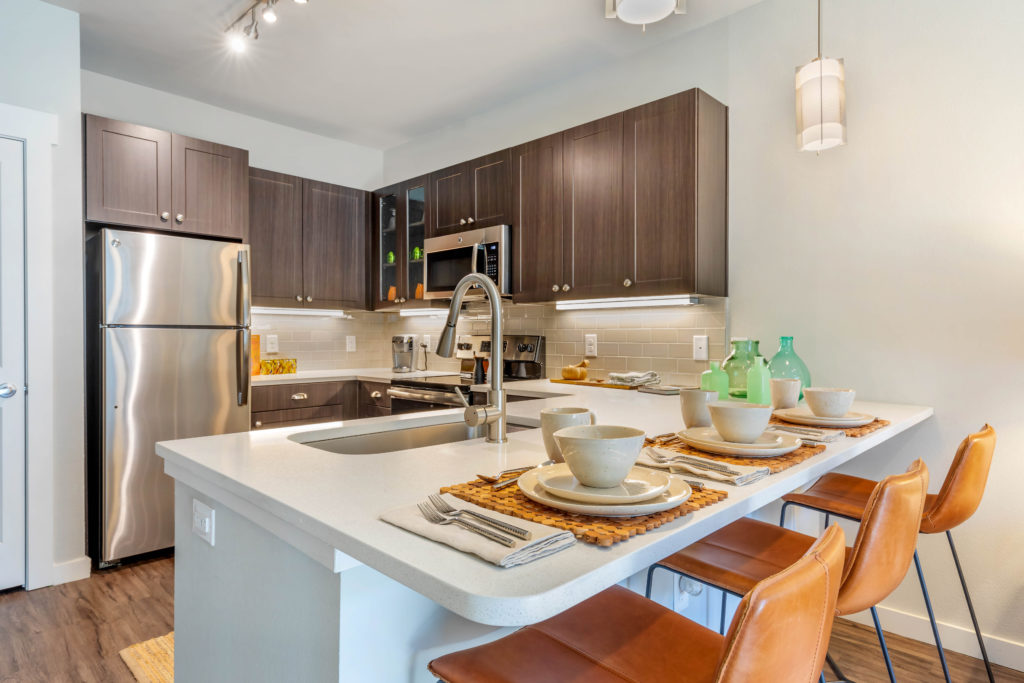 U-shaped kitchen with countertop bar seating 3 with place settings, large single basin sink with gooseneck faucet, dark wood upper and lower cabinetry and stainless steel appliances.
